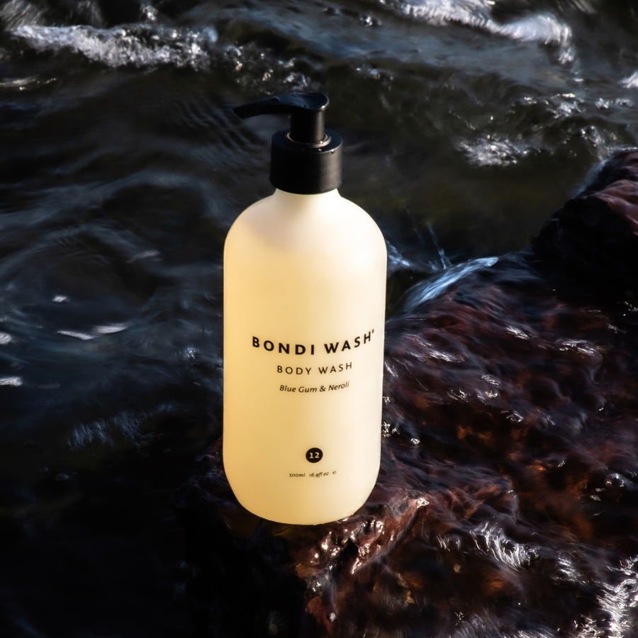A bottle of Bondi Wash body wash with a black pump dispenser, filled with a pale yellow liquid, set against a flowing dark stream.