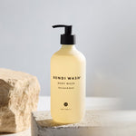 A bottle of Bondi Wash Body Wash with a black pump dispenser, filled with a pale yellow liquid, displayed on a textured stone pedestal next to a natural rock and white flowers.