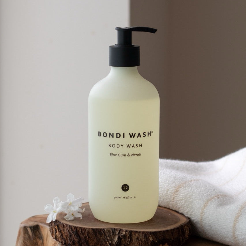 A bottle of Bondi Wash body wash with a black pump dispenser, resting on a rustic wooden slice with a white towel in the background.