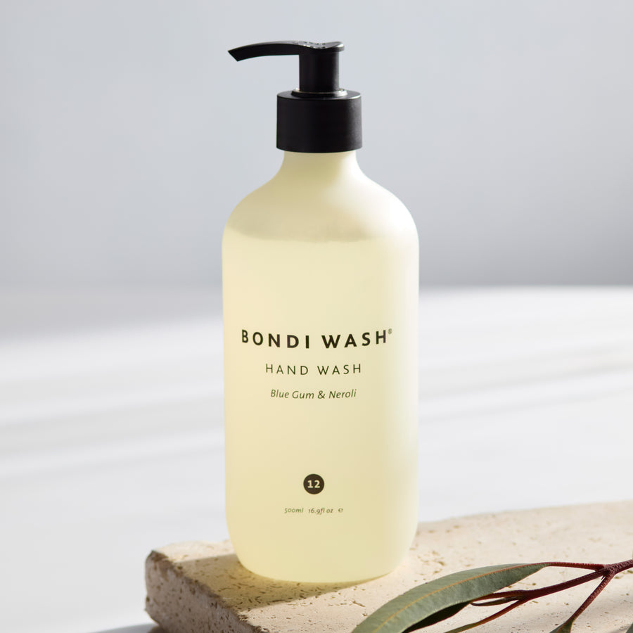 A bottle of Bondi Wash Blue Gum & Neroli hand wash, featuring a frosted pale yellow bottle with a black pump dispenser, styled on a textured stone slab with green leaves against a bright, clean background.