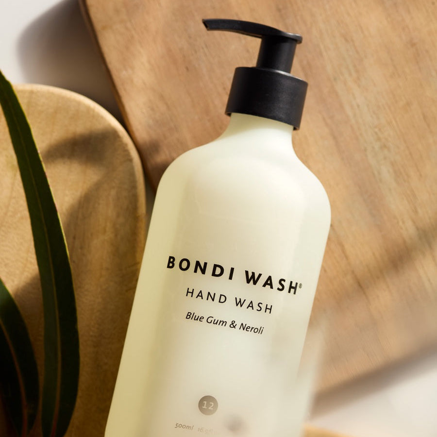 A frosted white pump bottle of Bondi Wash Hand Wash with Blue Gum and Neroli scent, styled on a wooden cutting board with a green leaf in the foreground.