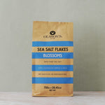A 750g bag of Olsson's Sea Salt Flakes Blossoms standing upright on a light marble surface against a textured gray wall.