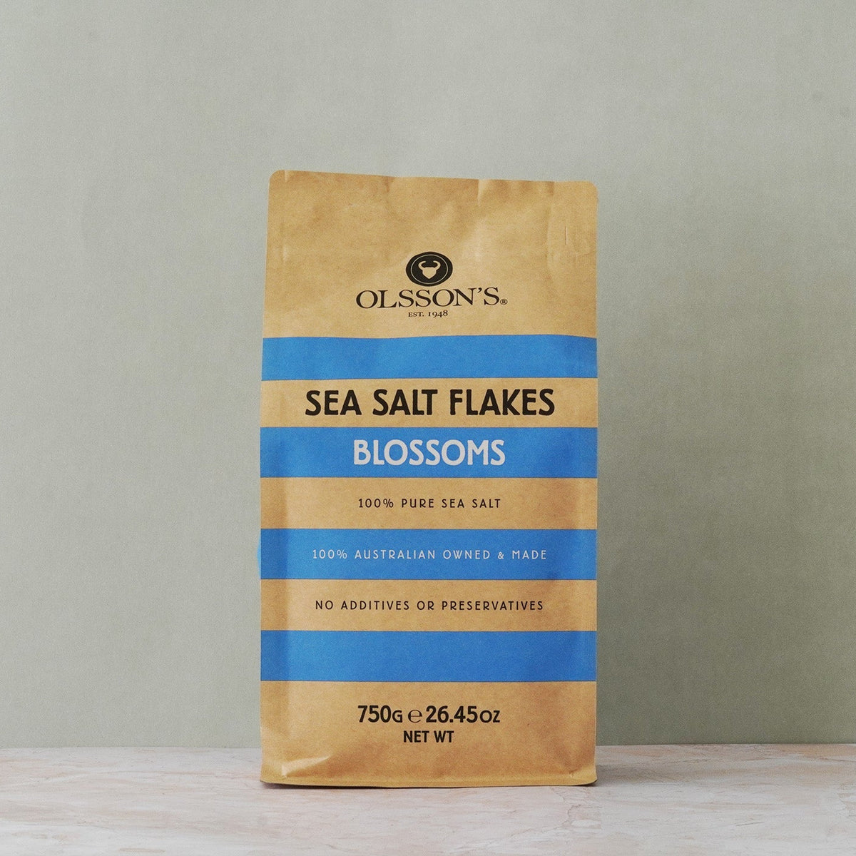 A 750g bag of Olsson's Sea Salt Flakes Blossoms standing upright on a light marble surface against a textured gray wall.