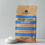 A brown paper bag of Olsson's Sea Salt Flakes Blossoms stands on a light surface against a pale gray wall, with a small white dish of salt in front of the package.