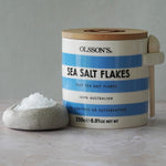 Container of Olsson's Sea Salt Flakes with a small bowl of salt on a neutral background