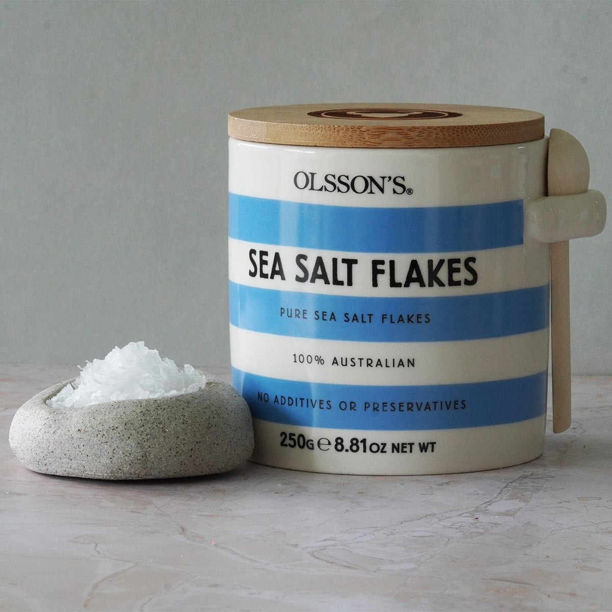 Container of Olsson's Sea Salt Flakes with a small bowl of salt on a neutral background