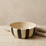 A handmade ceramic bowl with a cream-colored glaze and bold, hand-painted black vertical stripes, resting on a neutral beige surface with a matching cloth in the background.