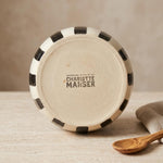 A round ceramic plate or dish shown from the bottom, featuring a cream-colored center with the brand name 'Charlotte Manser' printed in black, surrounded by a black and cream checkered rim. The plate is set on a light textured surface next to a wooden spoon and a folded cloth.