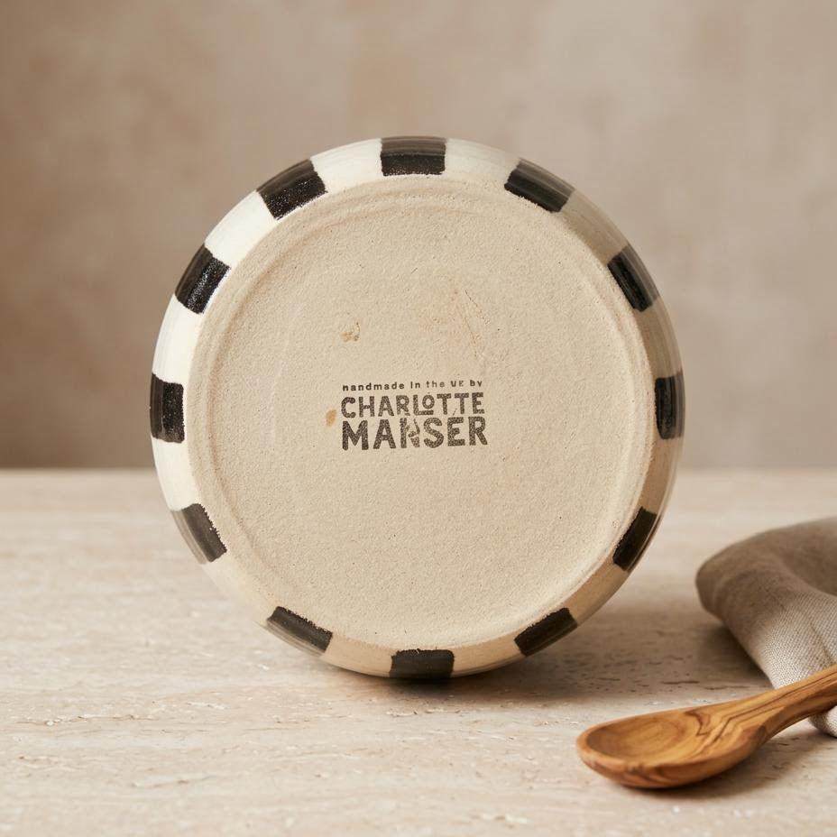 A round ceramic plate or dish shown from the bottom, featuring a cream-colored center with the brand name 'Charlotte Manser' printed in black, surrounded by a black and cream checkered rim. The plate is set on a light textured surface next to a wooden spoon and a folded cloth.