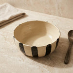 A handmade ceramic bowl with a cream-colored interior and exterior decorated with bold black vertical brushstrokes, styled on a beige stone surface next to a wooden spoon and a folded cloth.