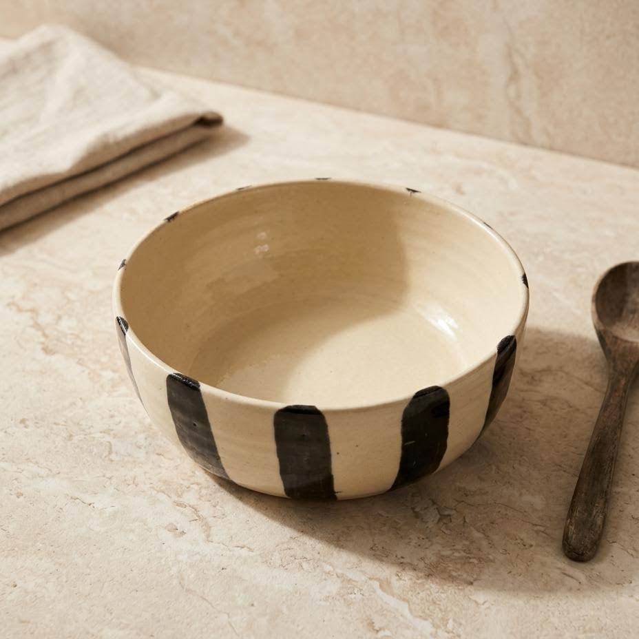A handmade ceramic bowl with a cream-colored interior and exterior decorated with bold black vertical brushstrokes, styled on a beige stone surface next to a wooden spoon and a folded cloth.