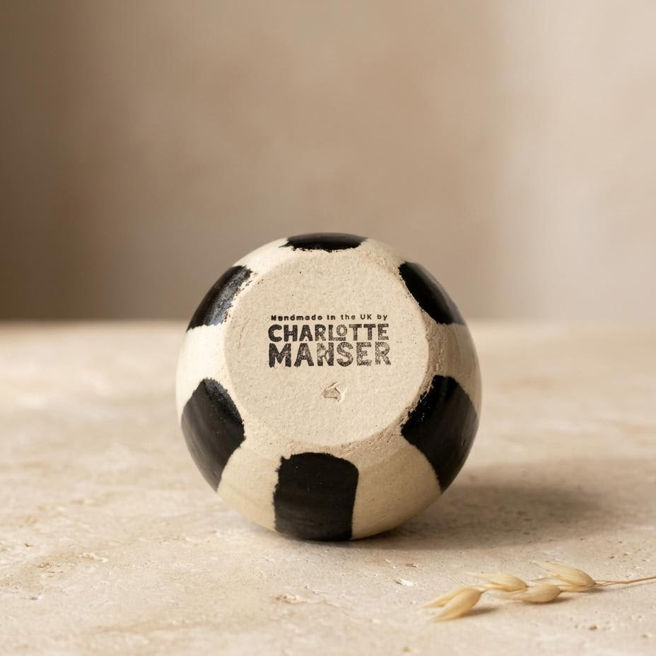 A small decorative bowl with a black and off-white stripe pattern sits on a textured beige surface. The center patch reads 'Handmade in the UK by CHARLOTTE MANSER' in black text.
