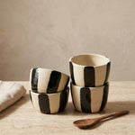 Four small ceramic cups with black and cream vertical stripes, stacked on a light wood surface next to a wooden spoon and a folded cloth, against a neutral beige background.
