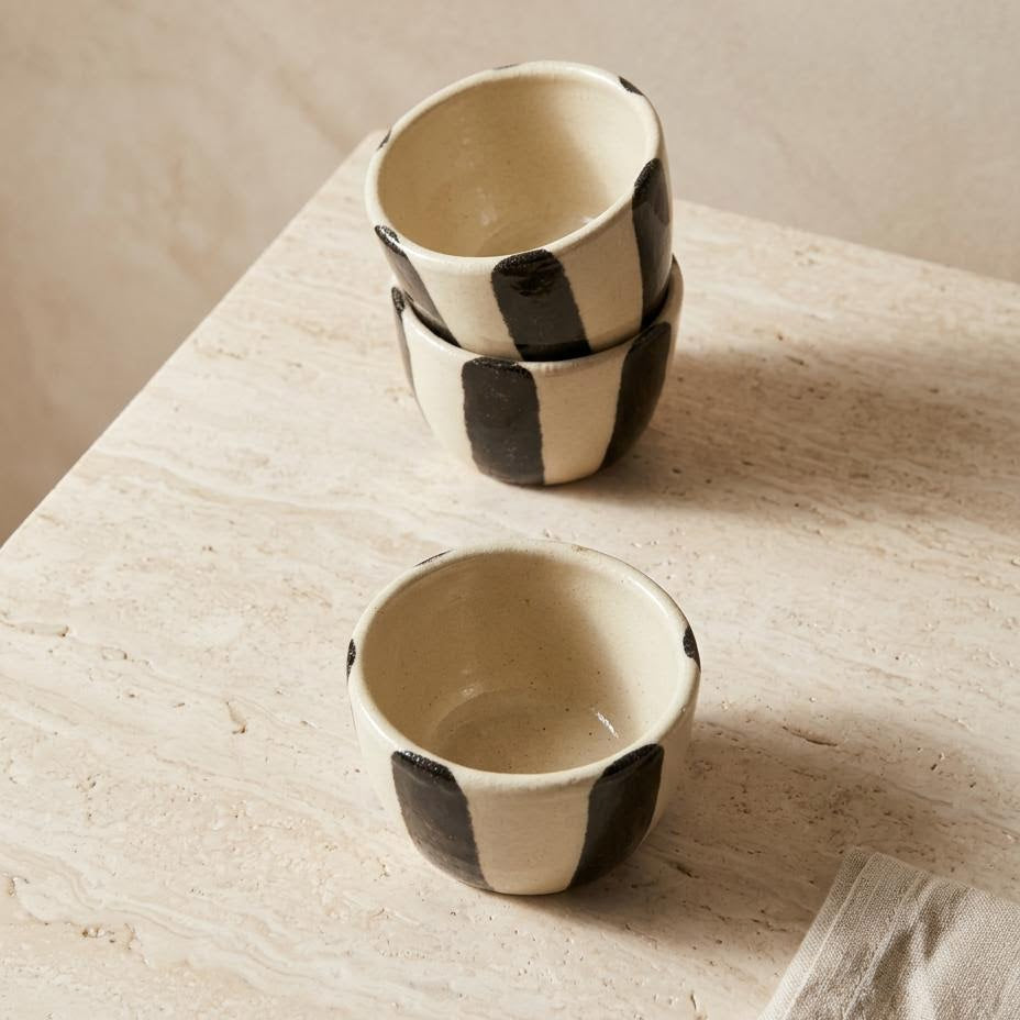 Two small handmade ceramic bowls with a black and cream checkerboard pattern, stacked on a light beige stone surface.