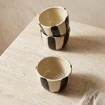Two small handmade ceramic bowls with a black and cream checkerboard pattern, stacked on a light beige stone surface.