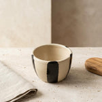 A small handmade ceramic bowl with a cream-colored glaze and bold black vertical stripes, styled on a textured stone surface with a folded linen napkin and a wooden lid nearby.