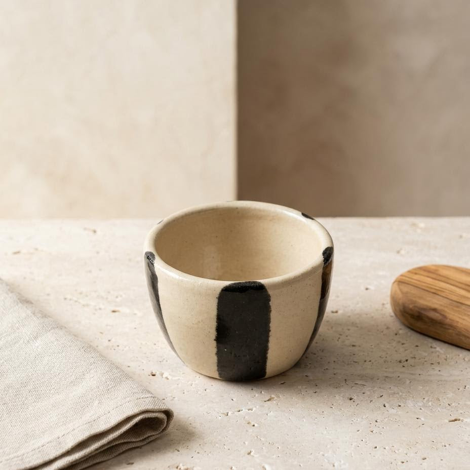 A small handmade ceramic bowl with a cream-colored glaze and bold black vertical stripes, styled on a textured stone surface with a folded linen napkin and a wooden lid nearby.
