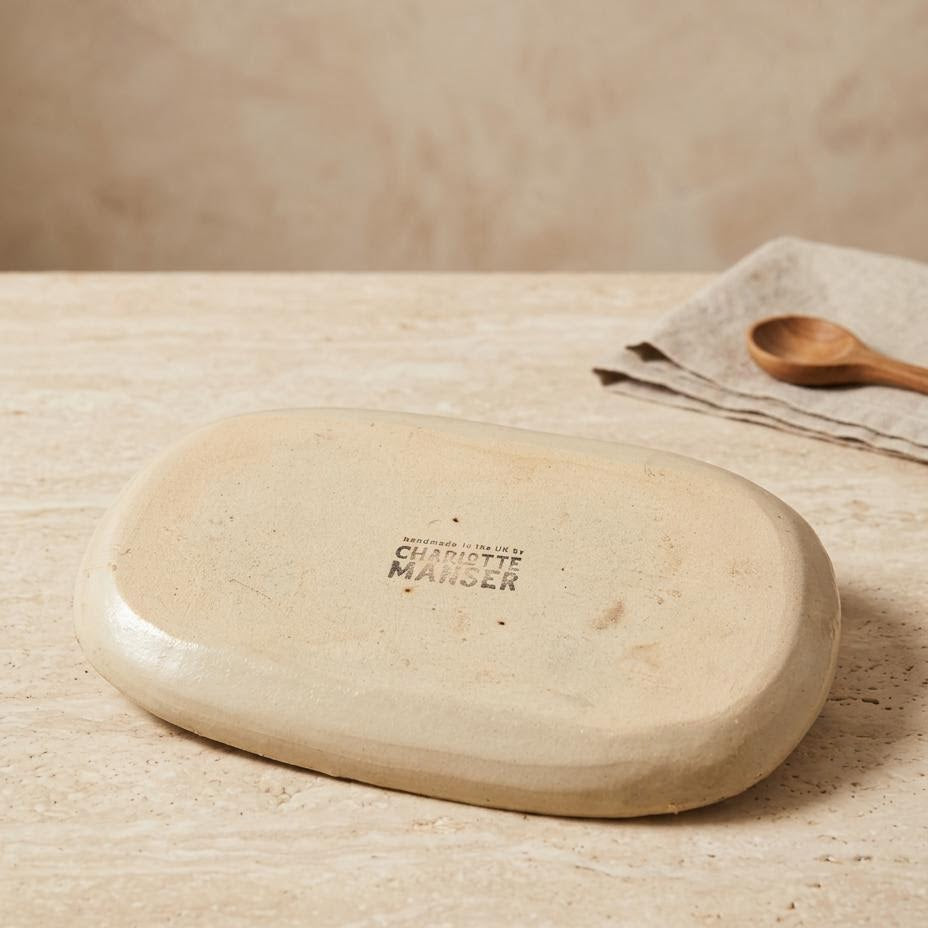 An oval ceramic dish or tray with a matte, warm beige finish, featuring a stamped brand logo reading 'CHARLOTTE MANSER'. It is displayed on a textured beige stone surface with a matching beige napkin and wooden spoon in the background.