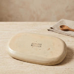 An oval ceramic dish or tray with a matte, warm beige finish, featuring a stamped brand logo reading 'CHARLOTTE MANSER'. It is displayed on a textured beige stone surface with a matching beige napkin and wooden spoon in the background.
