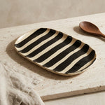 A rectangular ceramic serving platter with bold black and cream stripes, styled on a textured stone surface next to a wooden spoon and a beige linen cloth.