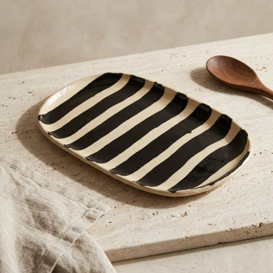 A rectangular ceramic serving platter with bold black and cream stripes, styled on a textured stone surface next to a wooden spoon and a beige linen cloth.