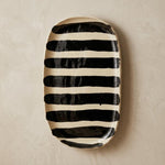 An oval ceramic serving platter with bold black hand-painted horizontal stripes, displayed on a textured beige wall above a styled tabletop with a napkin, wooden slice, and eucalyptus sprigs.