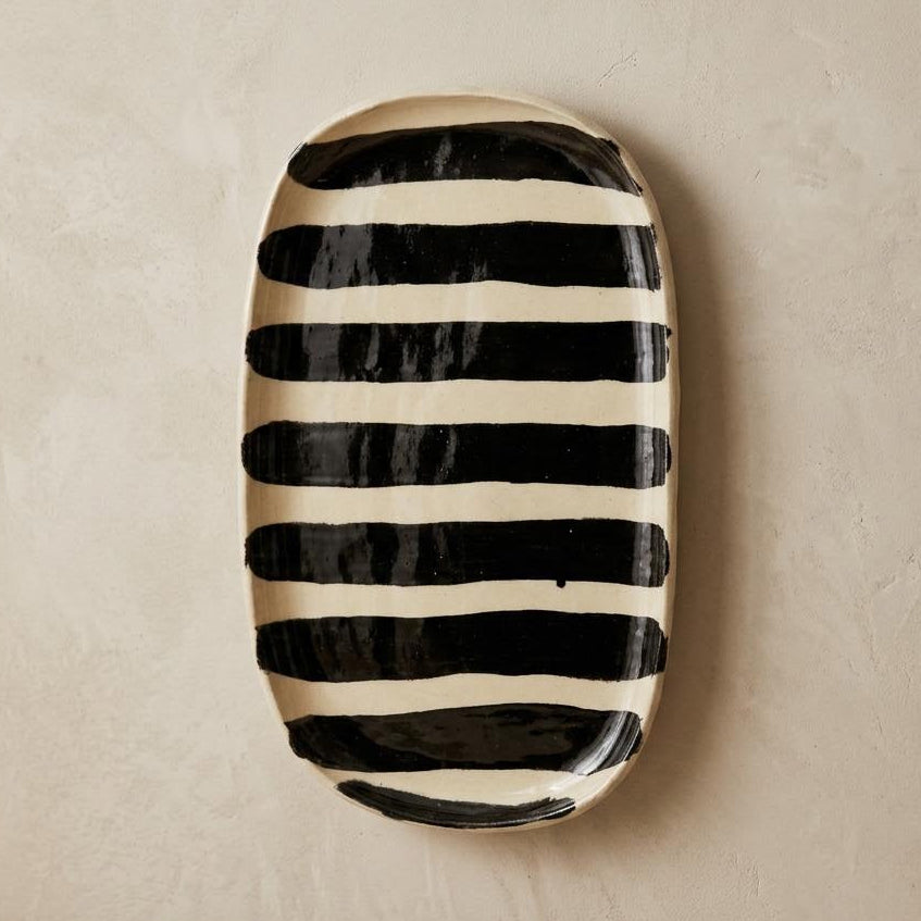 An oval ceramic serving platter with bold black hand-painted horizontal stripes, displayed on a textured beige wall above a styled tabletop with a napkin, wooden slice, and eucalyptus sprigs.