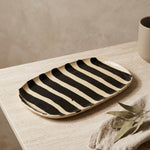 A rectangular ceramic serving platter with bold black and cream stripes, styled on a light beige stone table next to a linen napkin and dried leaves.