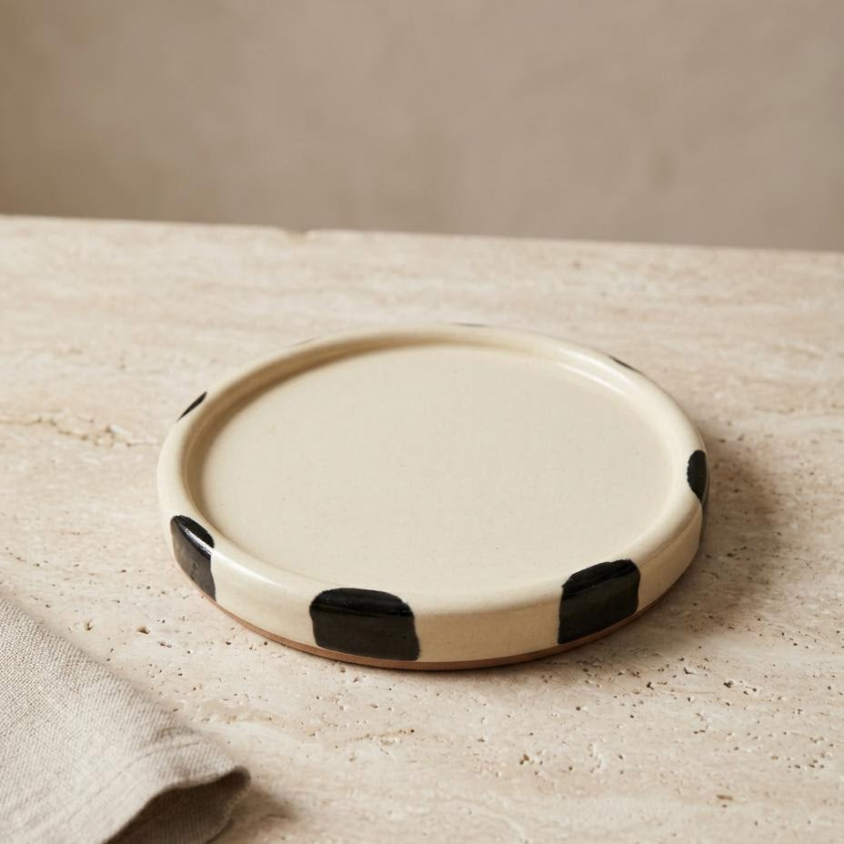 A round ceramic plate with a cream-colored glaze and black hand-painted abstract spots, resting on a textured beige stone surface next to a folded linen napkin.