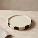 A round ceramic plate with a cream-colored glaze and black hand-painted abstract spots, resting on a textured beige stone surface next to a folded linen napkin.