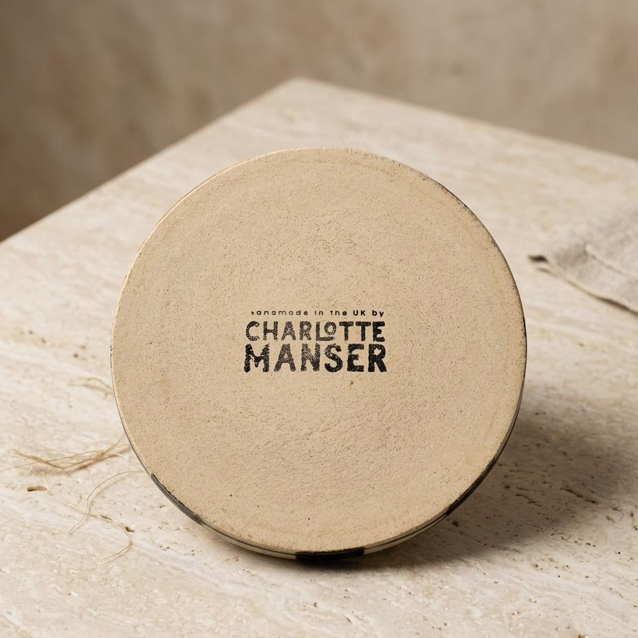 A round beige cosmetic compact lid with black text reading 'CHARLOTTE MANSER' and smaller text above, photographed on a light stone surface against a warm neutral background.