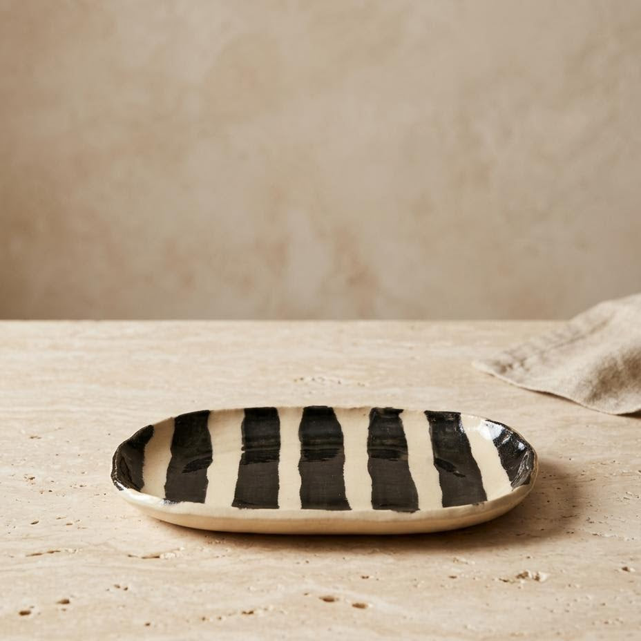 A small oval ceramic dish with bold black and cream vertical stripes, resting on a textured beige stone surface against a matching beige background.