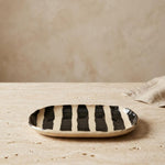 A small oval ceramic dish with bold black and cream vertical stripes, resting on a textured beige stone surface against a matching beige background.