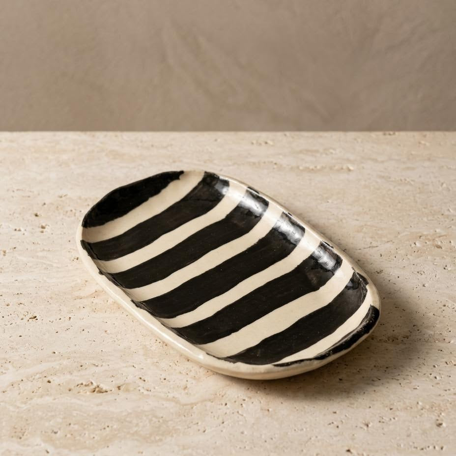 A small oval ceramic dish with bold black and cream horizontal stripes, resting on a textured beige stone surface against a neutral beige background.