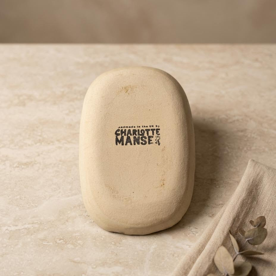A cream-colored ceramic soap dish with a black printed logo reading 'Charlotte Manse' on a textured beige stone surface, styled with a linen napkin and dried eucalyptus leaves.
