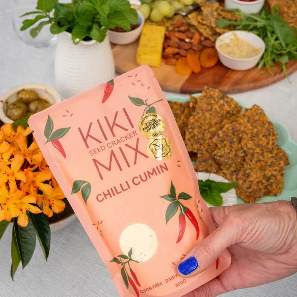 A hand holds a pink pouch of Kiki Seed Cracker Mix Chilli Cumin in front of a styled charcuterie board with crackers, cheese, nuts, and herbs on a white tablecloth.