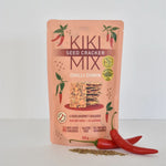 A stand-up pouch of Kiki Seed Cracker Mix Chilli Cumin crackers, shown on a clean white background with two red chili peppers and a small pile of crumbs in front. The packaging is peach-pink with green chili illustrations and a central image of the cracker slices.