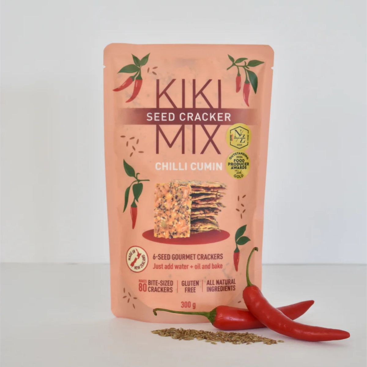 A stand-up pouch of Kiki Seed Cracker Mix Chilli Cumin crackers, shown on a clean white background with two red chili peppers and a small pile of crumbs in front. The packaging is peach-pink with green chili illustrations and a central image of the cracker slices.
