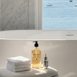 A bottle of KomiDai Wash Body Wash and a small dropper bottle with folded white towels, styled on a stone pedestal in front of a bathtub with a window overlooking the sea.