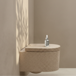 A wall-mounted toilet in a warm beige stone or concrete finish, shown in a minimalist bathroom setting with sunlight casting leaf shadows on the wall. A small glass dropper bottle sits on top of the toilet lid.