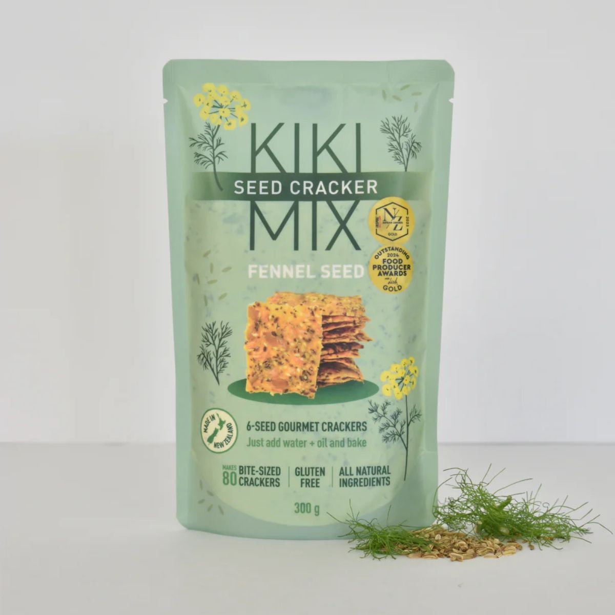 A 300g stand-up pouch of Kiki Seed Cracker Mix (Fennel Seed) on a white background, with a small pile of the crackers and fennel seeds in front of the package.