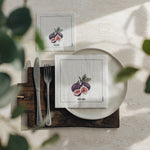 Two square white napkins with a purple fig illustration are arranged on a place setting. One napkin is on a white plate with a knife and fork on a dark wooden board, and the other is placed above on a light surface.