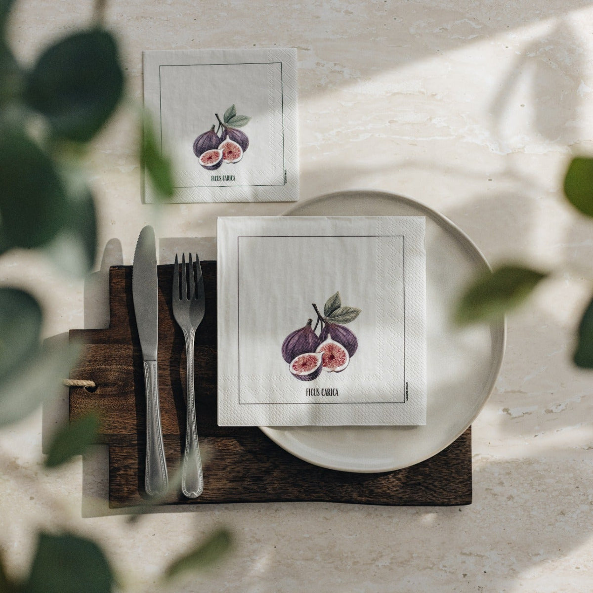 Two square white napkins with a purple fig illustration are arranged on a place setting. One napkin is on a white plate with a knife and fork on a dark wooden board, and the other is placed above on a light surface.