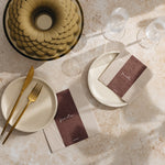 A styled tabletop flat lay with beige plates, gold-toned cutlery, clear drinking glasses, and matching brown-and-beige place cards on a light stone surface with soft shadows.