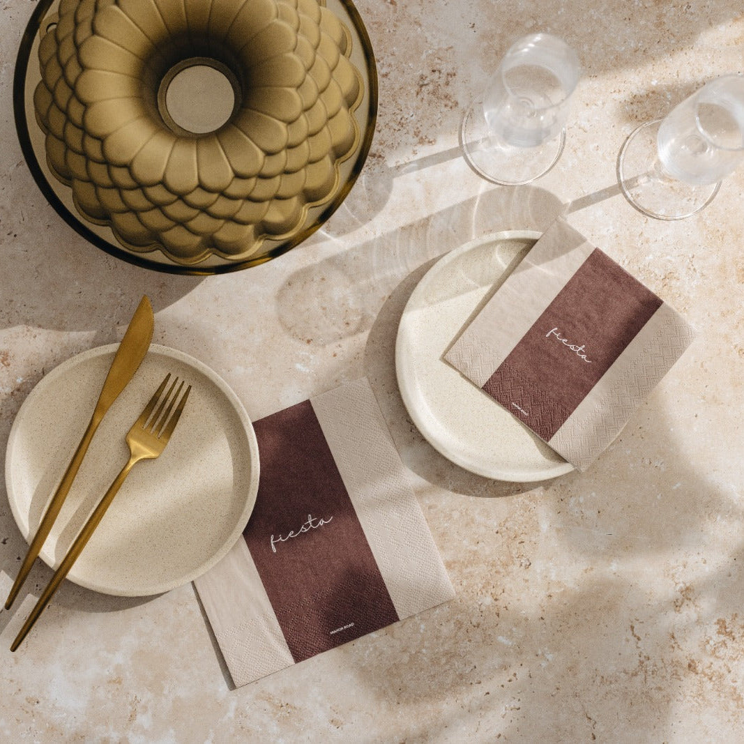 A styled tabletop flat lay with beige plates, gold-toned cutlery, clear drinking glasses, and matching brown-and-beige place cards on a light stone surface with soft shadows.