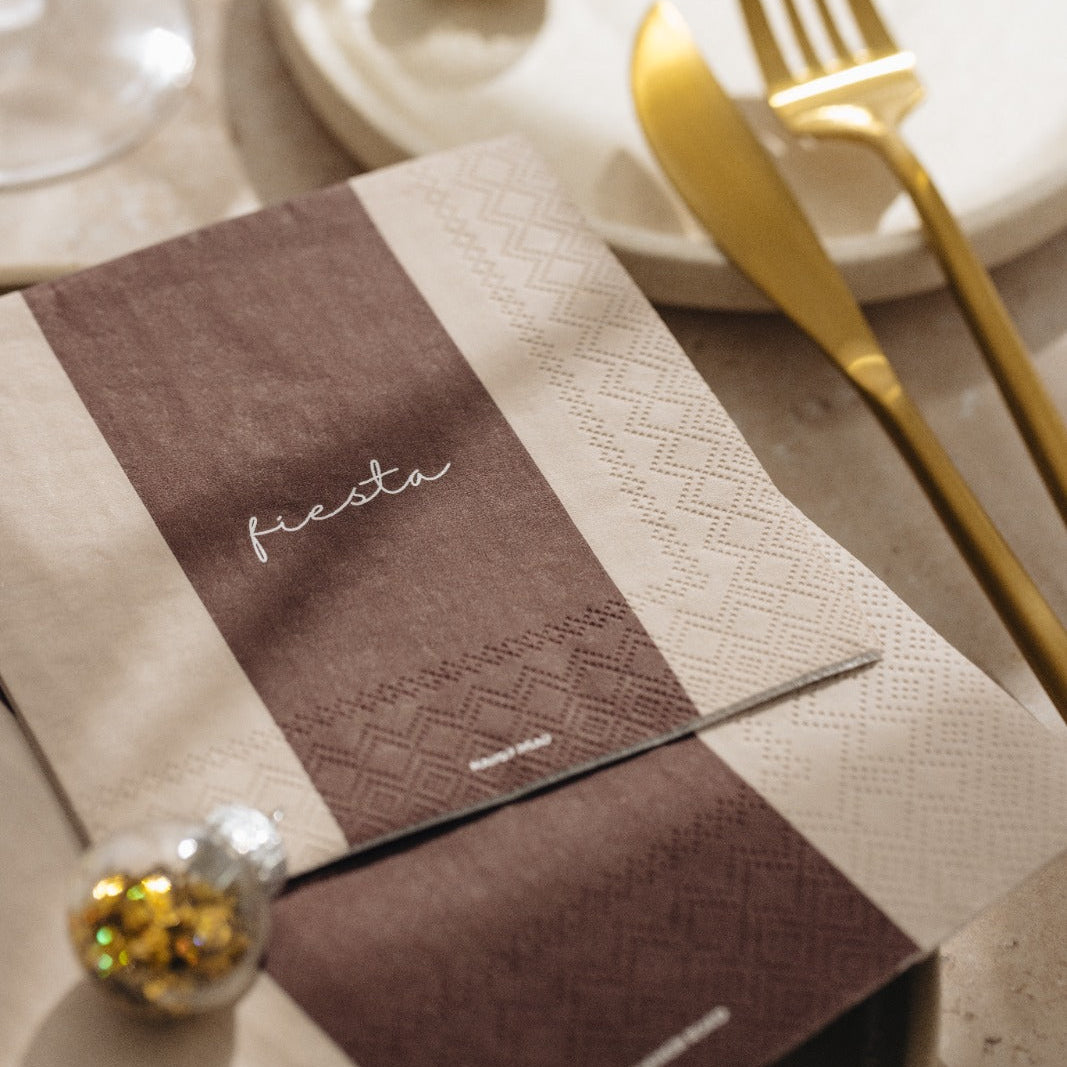 A styled place setting with a brown and beige patterned napkin featuring the word 'festa' in white script, accompanied by gold-toned flatware on a white plate and clear glassware in a softly lit tabletop setting.