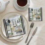 Two decorative napkins featuring a watercolor-style street cafe scene with black-and-white striped accents, styled on a beige table setting with a plate, fork, knife, teapot, and cup of tea.