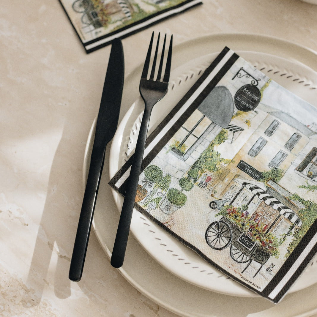 A styled tabletop scene with a white teacup and saucer, a white teapot, black flatware, and printed paper napkins featuring a vintage street-cafe illustration on a light stone surface.
