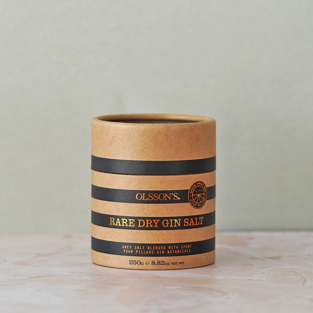 Olsson's Rare Dry Gin Salt in a cylindrical kraft paper container with black and gold stripes, set on a light marble surface against a neutral gray-green background.