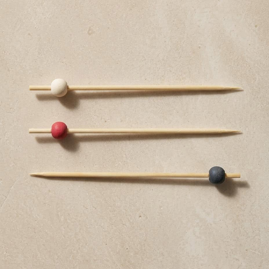 Three wooden toothpick-style picks with small round beads in white, red, and dark blue, arranged horizontally on a light beige textured background.
